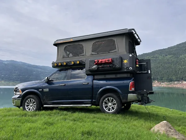 Blue RAM with a SUNHOMEE slide-in camper, roof raised beside a mountain lake